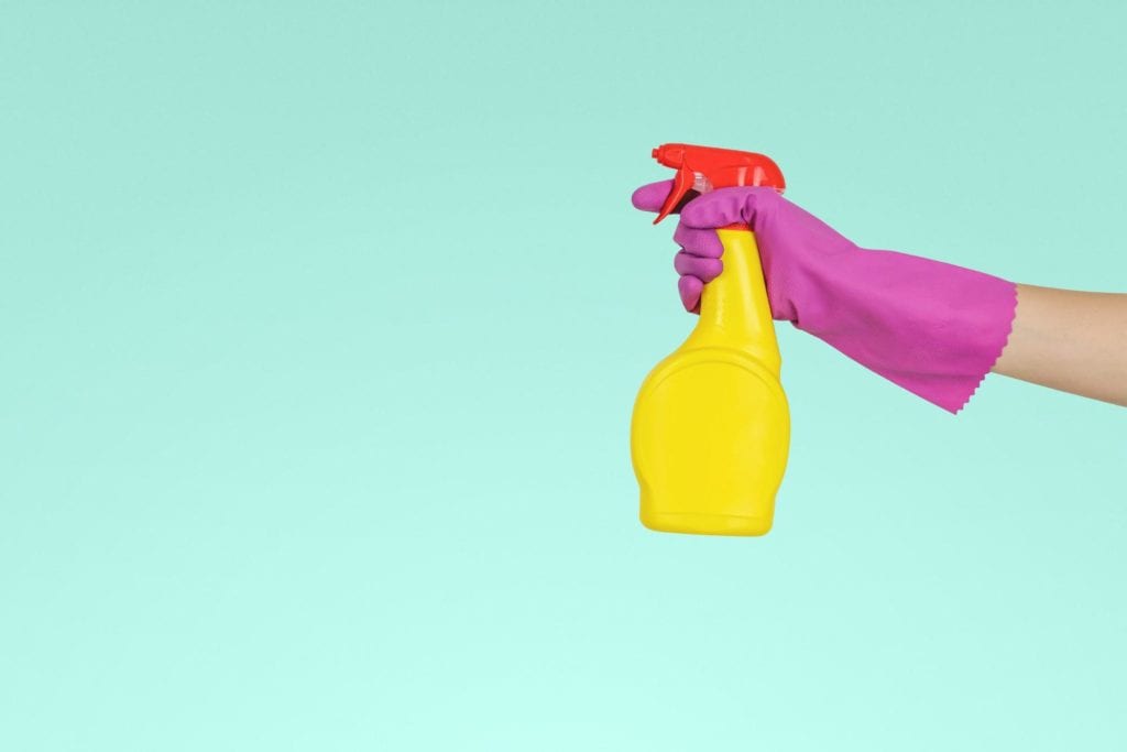 hand with pink cleaning glove holding yellow spray bottle in front of turquoise wall