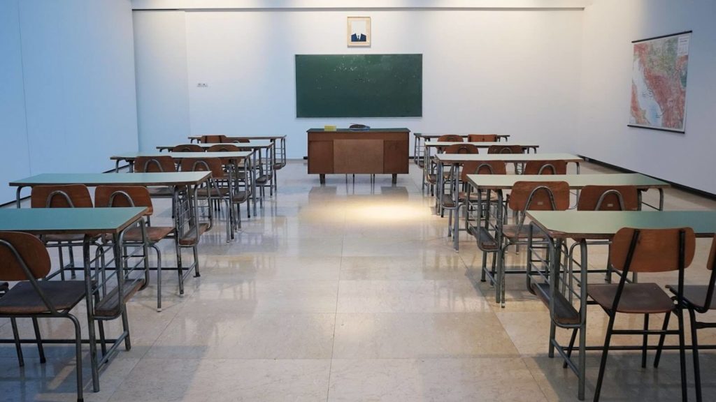 two rows of green desks with chairs in front of main desk with green chalkboard