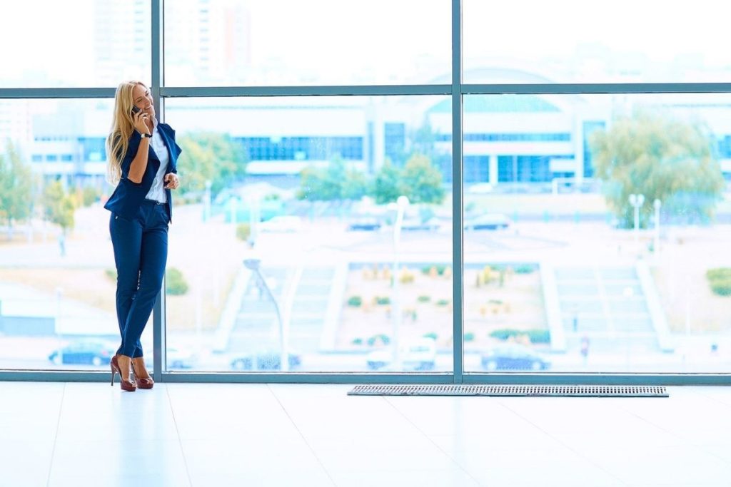 blonde woman in business suit talking on the phone leaning against big windows of building